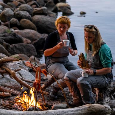 To kvinder tilbereder fisk over bål ved kysten