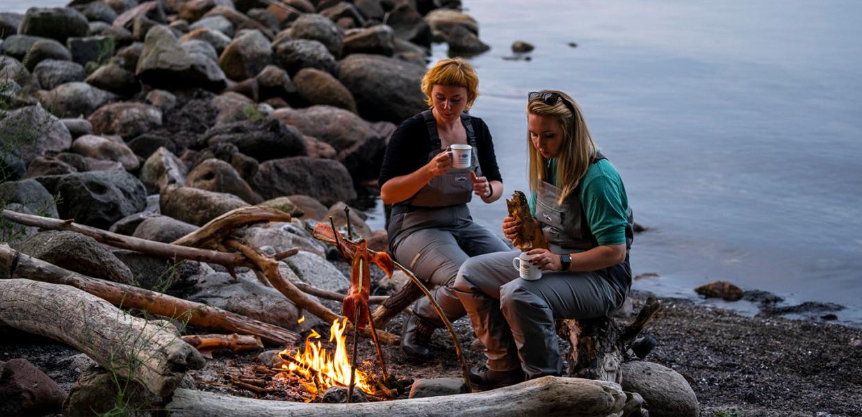 To kvinder tilbereder fisk over bål ved kysten